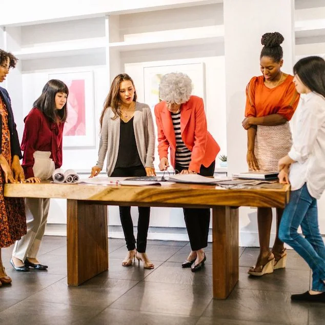 Six people stand around a wooden table, reviewing documents and discussing something together in a modern, well-lit office space with framed art on the walls. Among them are first-gen women, bringing unique perspectives to their collaborative approach as they explore how artificial intelligence can drive innovation within their team.